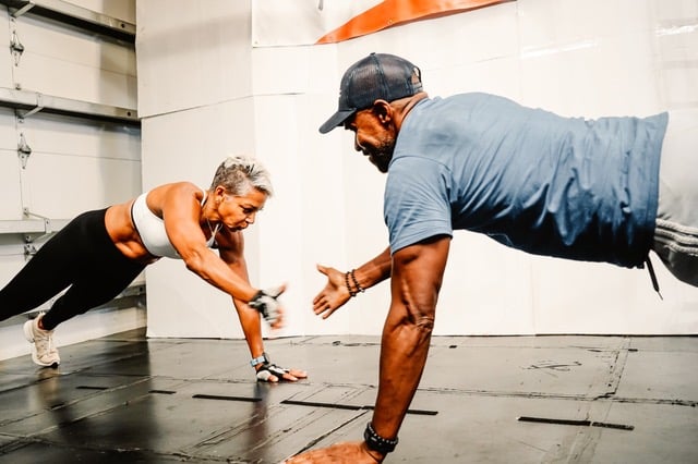 Nate and Shebah performing a dynamic plank exercise in a gym, reaching out for a high-five while maintaining proper form, showcasing strength and teamwork.