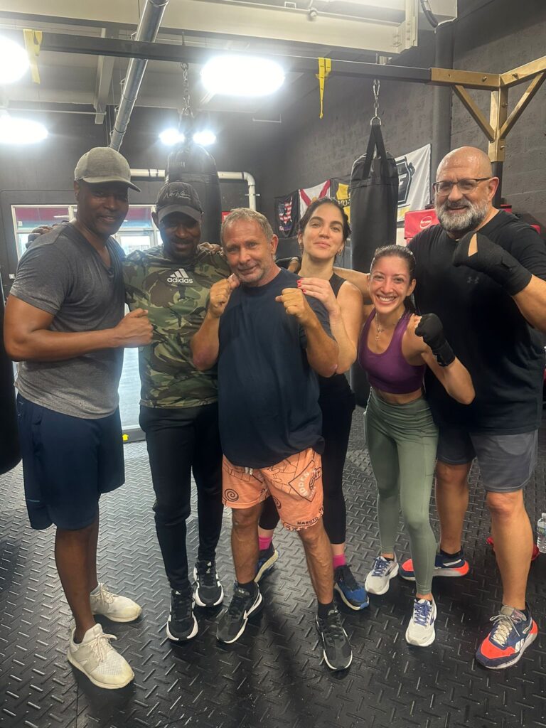 A group of diverse adults posing confidently in a boxing gym, showcasing camaraderie and strength, with punching bags and gym equipment in the background.