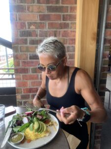 A woman with short gray hair, sunglasses, and a black tank top is enjoying a healthy meal at an outdoor cafe. Her plate contains a fresh salad with avocado slices, radishes, leafy greens, and an omelet.