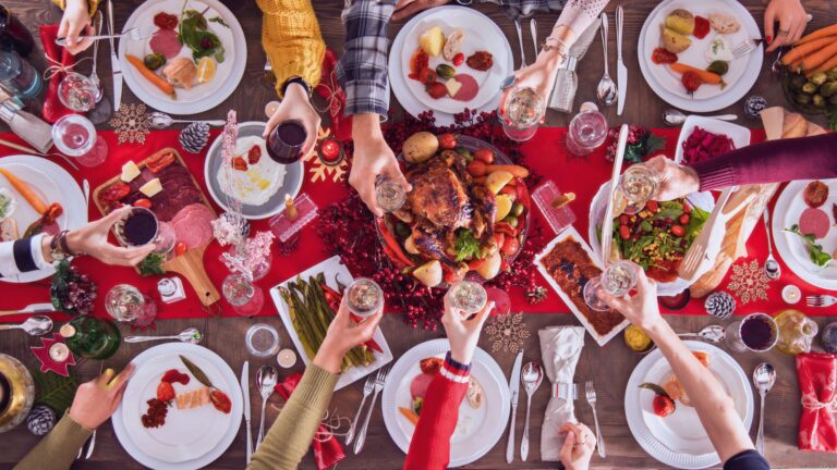 A festive holiday table viewed from above, filled with colorful dishes like roasted vegetables, fresh salads, lean proteins, and garnished platters. People are raising glasses in a toast, surrounded by holiday decorations, including red tablecloths, pinecones, and candles. Healthy Holiday Eating