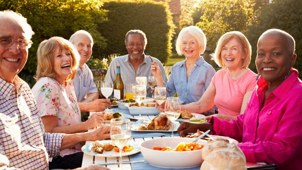 A group of vibrant, active seniors gathered around an outdoor dining table, laughing and enjoying a meal together in the warm sunlight.