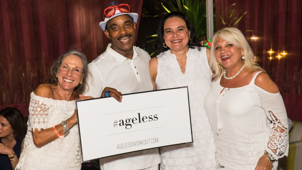 A group of four vibrant seniors in white outfits smiling and holding a sign that says "#ageless" at a social event by Nate Wilkins and Ageless Workout