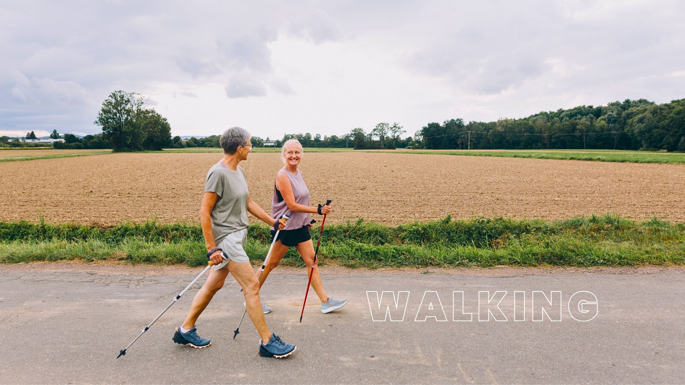 Two older women enjoying Nordic walking on a rural path, using walking poles for support, surrounded by open fields and greenery under a cloudy sky.