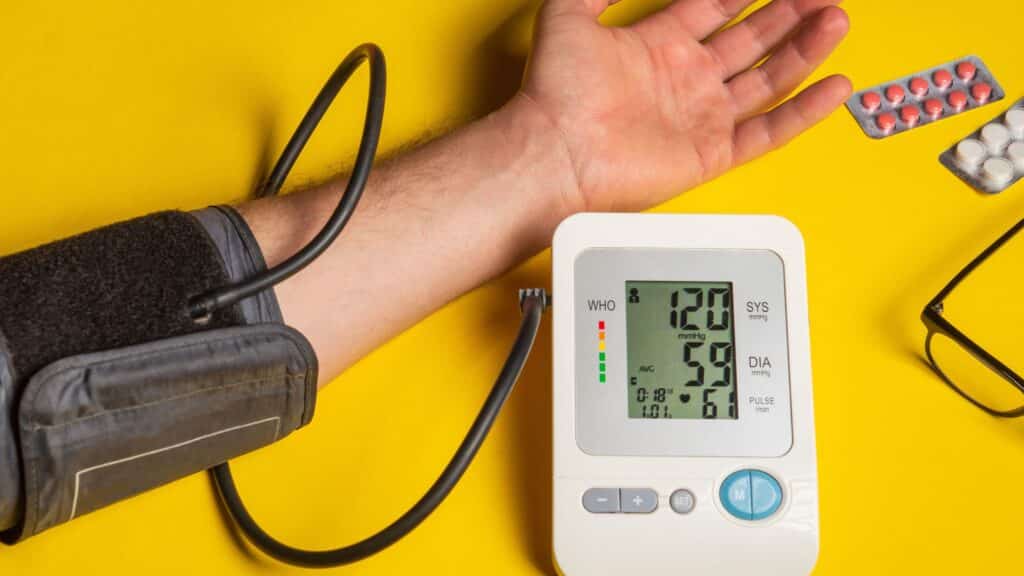A person checking their blood pressure with a digital monitor; the display shows a reading of 120/59 mmHg with a pulse of 61 bpm, set against a bright yellow background with pills and eyeglasses nearby.