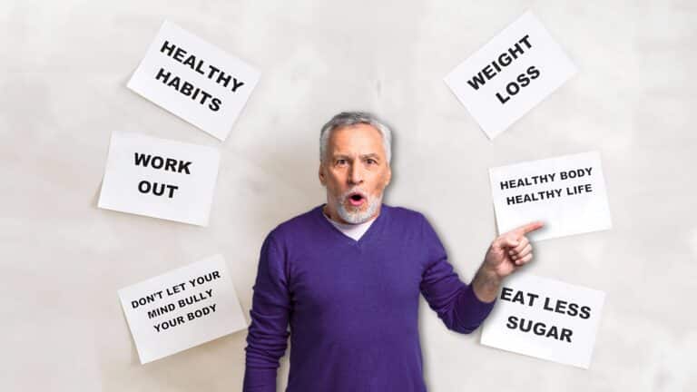 Older man in a purple sweater pointing to a sign that reads "Healthy Body Healthy Life," surrounded by motivational health and fitness posters.