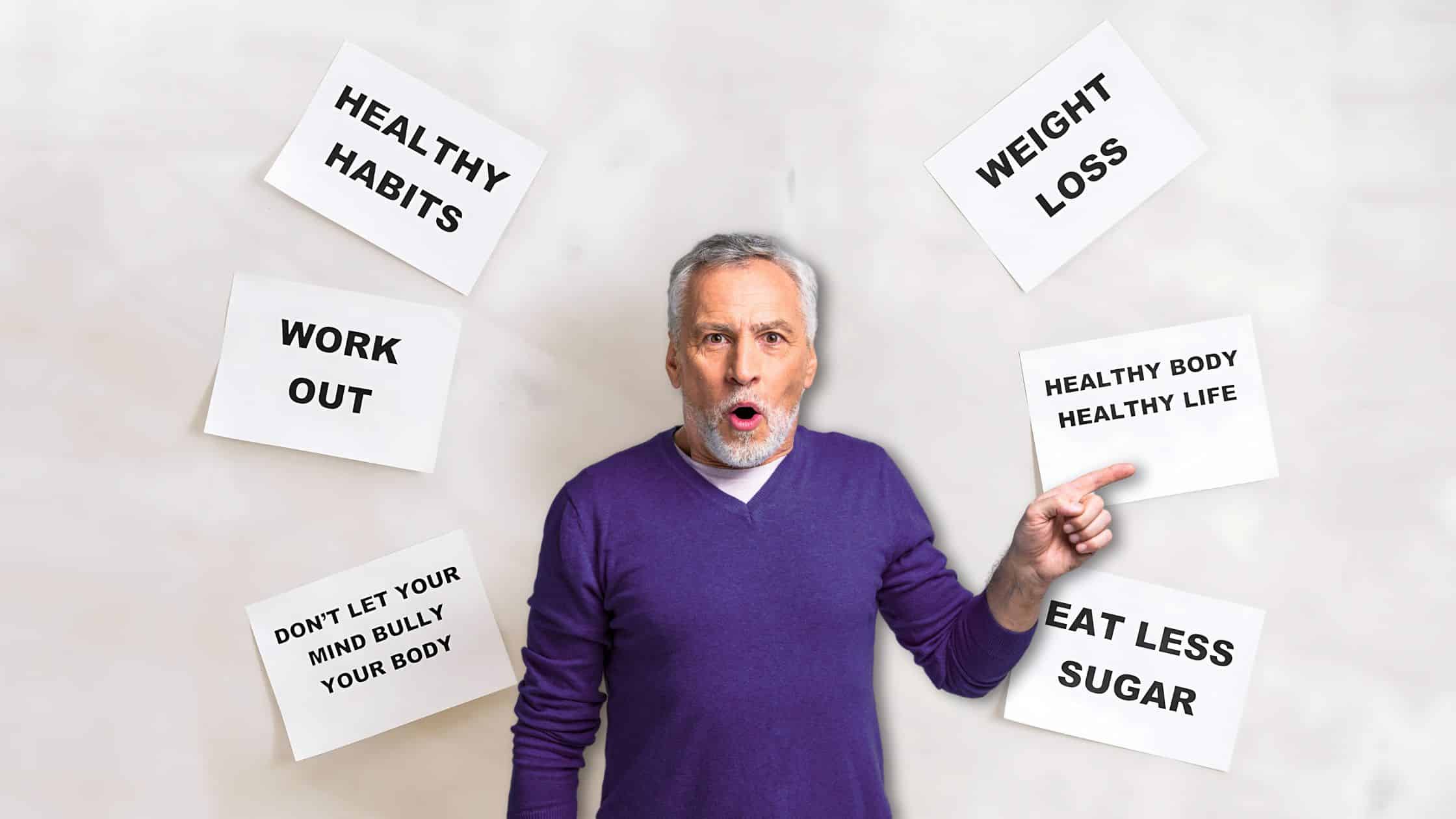 Older man in a purple sweater pointing to a sign that reads "Healthy Body Healthy Life," surrounded by motivational health and fitness posters.