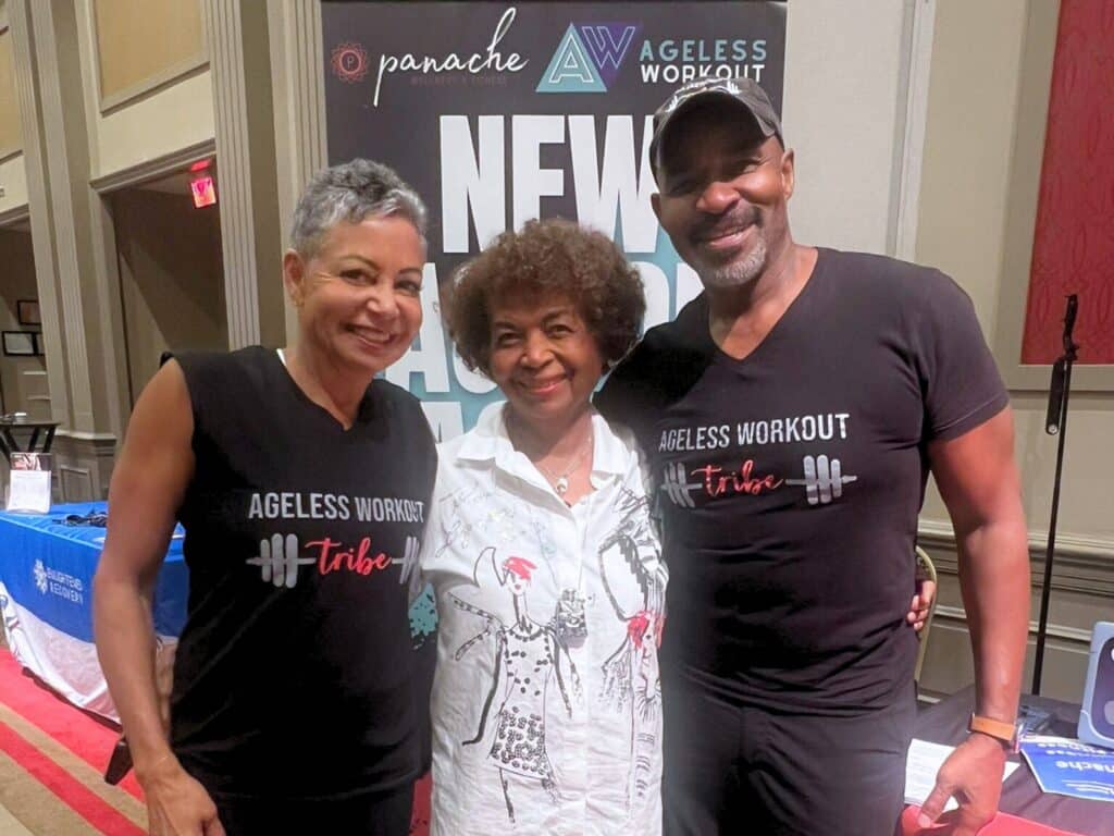 Three older adults stand together smiling, wearing athletic clothing; two wear black "Ageless Workout Tribe" shirts and the man in the center wears a black cap. AgelessWorkout Method showing active adults embracing healthy aging and vitality.