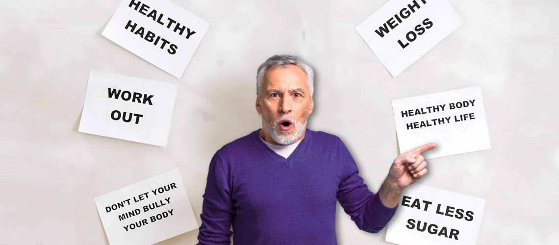 Older man in a purple sweater pointing to a sign that reads "Healthy Body Healthy Life," surrounded by motivational health and fitness posters.