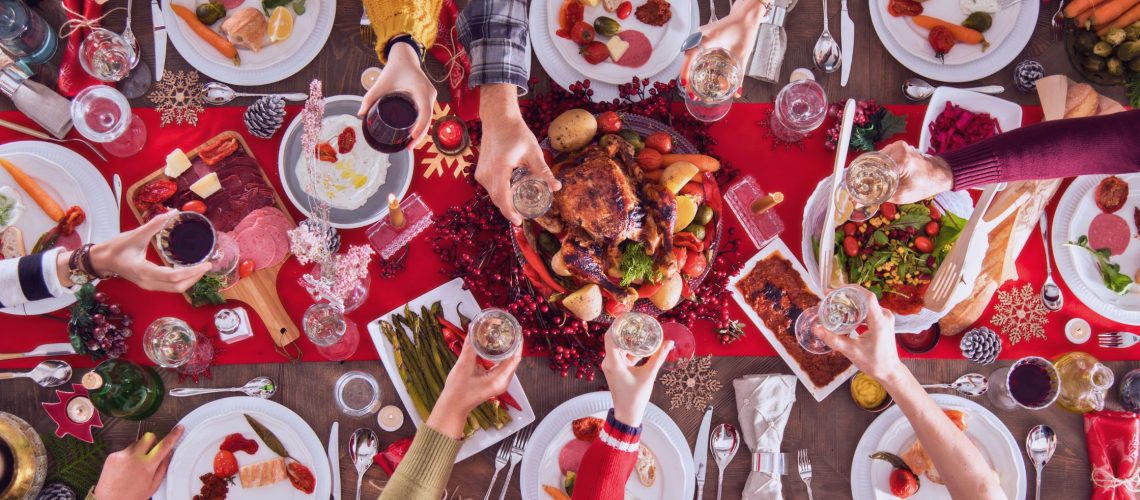 A festive holiday table viewed from above, filled with colorful dishes like roasted vegetables, fresh salads, lean proteins, and garnished platters. People are raising glasses in a toast, surrounded by holiday decorations, including red tablecloths, pinecones, and candles. Healthy Holiday Eating